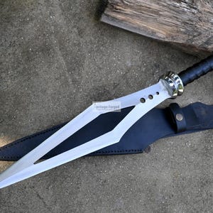Puede incluir: Una espada plateada con mango negro y una vaina de cuero. La espada tiene una hoja larga y puntiaguda con un dise&ntilde;o en forma de diamante. La vaina es de cuero negro con una correa de cuero marr&oacute;n.