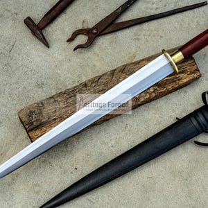 May include: A handmade dagger with a polished silver blade, a dark wood handle, and a brass guard. The dagger rests on a wooden plank, accompanied by a black leather sheath. Blacksmith tools are in the background.