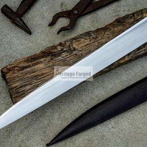 May include: A long, silver blade with a wooden handle and a leather sheath. The blade is sharp and pointed, and the handle is decorated with a metal design. The sheath is brown and has a flap that closes with a buckle.