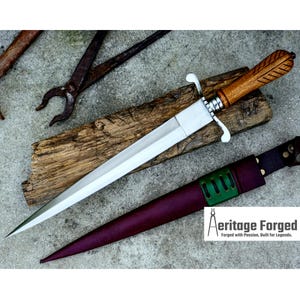 May include: A handmade dagger with a polished steel blade and a wooden handle with a leaf design. The dagger rests on a piece of wood, with a leather sheath in burgundy and green. The text "Aeritage Forged" is visible.