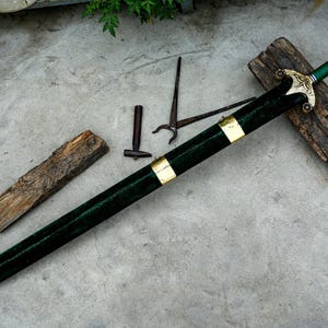 May include: A decorative sword with a green handle and scabbard, accented with gold-colored metal. The sword rests on a piece of wood, with other tools nearby. The blade is sheathed in a dark green material.