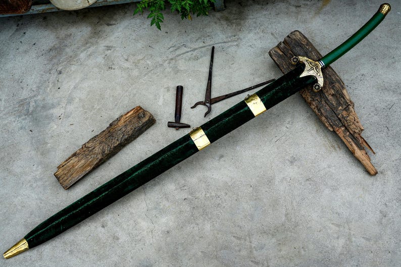 May include: A decorative sword with a green handle and scabbard, accented with gold-colored metal. The sword rests on a piece of wood, with other tools nearby. The blade is sheathed in a dark green material.