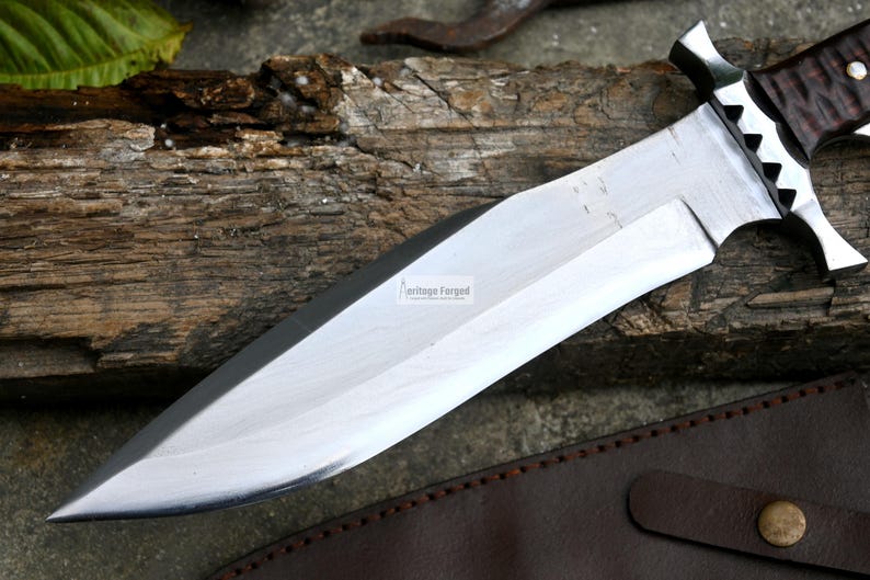 Puede incluir: Un cuchillo de caza plateado con mango de cuero marr&oacute;n y filo dentado. El cuchillo est&aacute; apoyado en una pieza de madera con una funda de cuero marr&oacute;n.