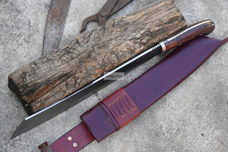 May include: A large, handmade knife with a dark wooden handle and a long, straight blade. The knife rests on a weathered wooden block, next to a burgundy leather sheath. The sheath has a brown leather belt loop. The knife is a tool.