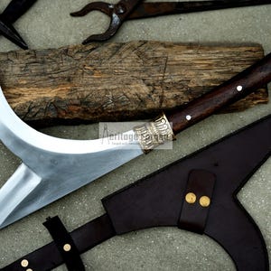 May include: A curved, silver-colored blade with a brown wooden handle and a decorative silver metal pommel. The blade is attached to a brown leather sheath with a strap.