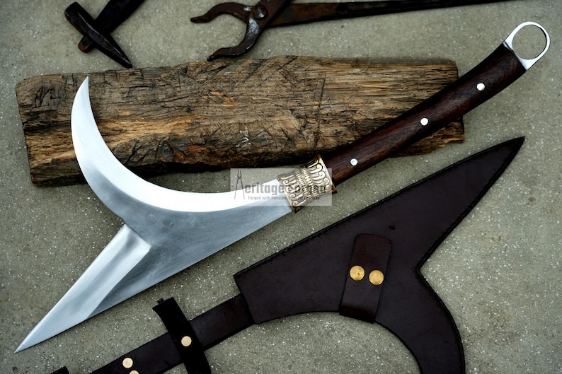 May include: A curved, silver-colored blade with a brown wooden handle and a decorative silver metal pommel. The blade is attached to a brown leather sheath with a strap.