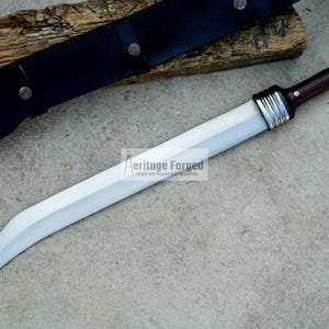 May include: A silver-colored sword with a brown wooden handle and a black leather sheath. The sword has a curved blade and a silver metal band around the handle. The text "Heritage Forged" is visible on the blade.