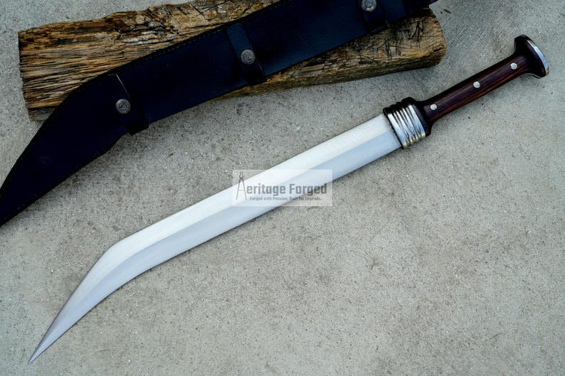 May include: A silver-colored sword with a brown wooden handle and a black leather sheath. The sword has a curved blade and a silver metal band around the handle. The text "Heritage Forged" is visible on the blade.