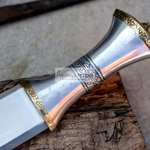 May include: A silver dagger with a gold-colored handle featuring intricate floral designs. The handle is shaped like a bell and has a pointed tip.