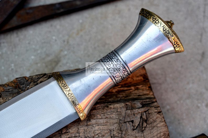May include: A silver dagger with a gold-colored handle featuring intricate floral designs. The handle is shaped like a bell and has a pointed tip.
