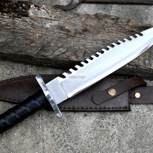 Pode incluir: Uma faca de prata com cabo preto e l&acirc;mina serrilhada. A faca tem uma bainha de couro com um bot&atilde;o de lat&atilde;o. A faca &eacute; etiquetada como "Handmade Forged".