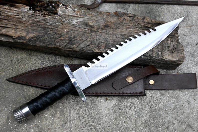 Pode incluir: Uma faca de prata com cabo preto e l&acirc;mina serrilhada. A faca tem uma bainha de couro com um bot&atilde;o de lat&atilde;o. A faca &eacute; etiquetada como "Handmade Forged".