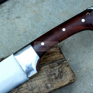 May include: A close-up of a silver knife with a dark wood handle. The handle has two white dots and the text "Heritage Forged" is visible on the blade.
