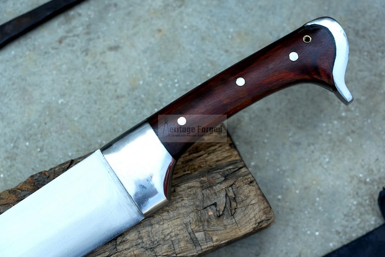 May include: A close-up of a silver knife with a dark wood handle. The handle has two white dots and the text "Heritage Forged" is visible on the blade.