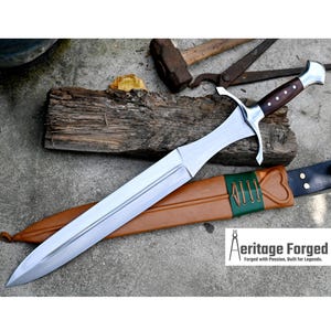 Könnte beinhalten: Ein silbernes Schwert mit einem braunen Holzgriff und einer grünen Lederscheide. Das Schwert liegt auf einer Holzoberfläche mit einem Hammer und einer Zange im Hintergrund. Der Text "Heritage Forged" ist in der unteren rechten Ecke des Bildes sichtbar. "Forged with Passion, Built for Legends."