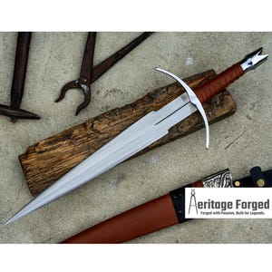 May include: A silver sword with a brown leather-wrapped handle and a cross-shaped guard rests on a wooden plank. The sword is next to a brown leather sheath. The image also shows blacksmith tools.
