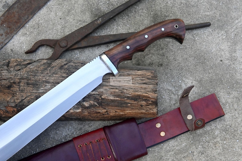 May include: A large, polished steel blade knife with a dark wood handle and silver accents. The knife rests on a wooden surface, accompanied by a leather sheath and blacksmith tools. The knife has a curved handle and a wide blade.