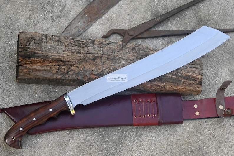 May include: A large, silver-bladed machete with a dark wood handle and a leather sheath. The blade is resting on a piece of wood, with other tools in the background. The sheath is burgundy in color.