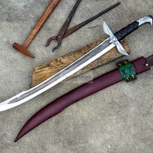 May include: A handcrafted sword with a silver blade and black handle, resting on a wooden plank. A burgundy leather sheath with green accents and a hammer and pliers are also visible, suggesting a blacksmithing theme.