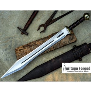 Könnte beinhalten: Ein silbernes Schwert mit einem braunen Ledergriff und einem goldenen Knauf. Das Schwert liegt auf einem Holzbrett und eine braune Lederscheide befindet sich im Hintergrund. Der Text "Heritage Forged" ist in der unteren rechten Ecke des Bildes sichtbar.  Forged with Passion, Built for Legends.