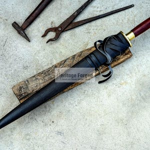 May include: A black dagger with a leather-wrapped handle and a wooden grip, resting on a wooden plank. The dagger has a dark blade and a gold-colored hilt. Black leather straps are wrapped around the handle. A hammer and pliers are in the background.