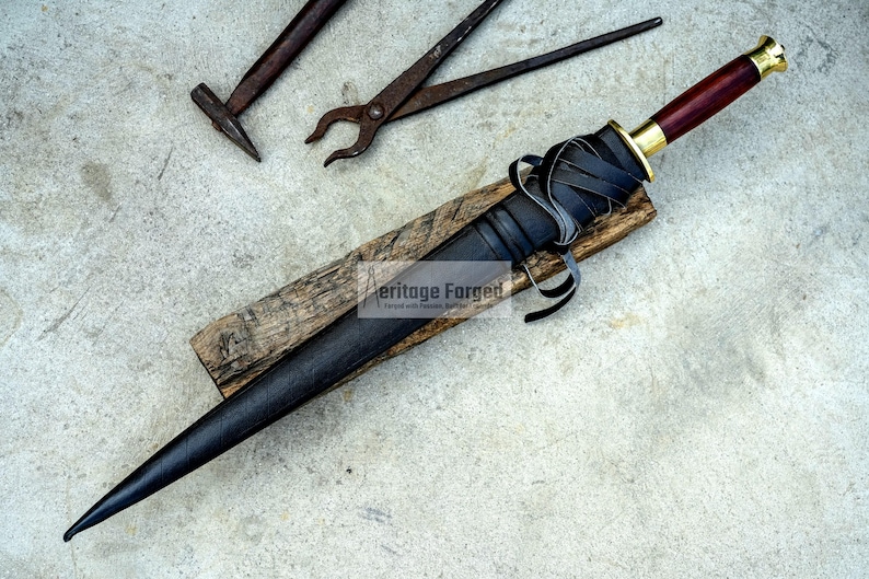 May include: A black dagger with a leather-wrapped handle and a wooden grip, resting on a wooden plank. The dagger has a dark blade and a gold-colored hilt. Black leather straps are wrapped around the handle. A hammer and pliers are in the background.