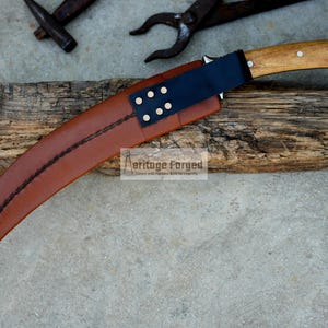 May include: A curved brown leather sheath with a black leather strap and brass rivets. The sheath is holding a curved knife with a wooden handle.