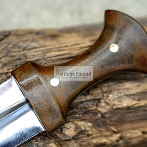 May include: A close-up of the handle of a knife with a dark brown wooden handle and a silver blade. The handle has a brass accent and the text "Heritage Forged" is visible on the handle.