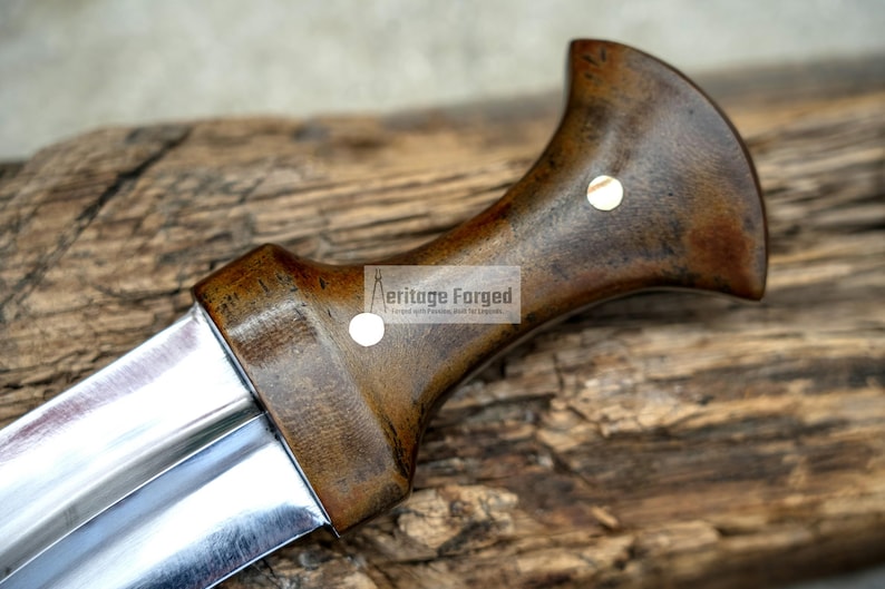 May include: A close-up of the handle of a knife with a dark brown wooden handle and a silver blade. The handle has a brass accent and the text "Heritage Forged" is visible on the handle.