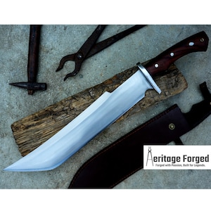 May include: A large, curved, silver knife with a wooden handle. The knife is resting on a wooden board and has a leather sheath. The text "Heritage Forged" is visible in the lower right corner of the image.  Forged with Passion, Built for Legends.