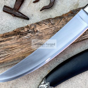 May include: A curved, silver knife with a black handle and a leather sheath. The knife is resting on a piece of weathered wood. The text "Heritage Forged" is visible on the blade.