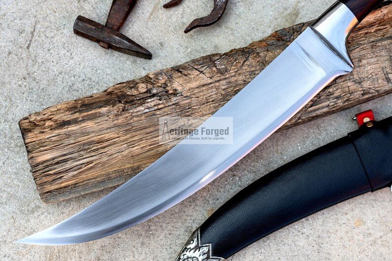 May include: A curved, silver knife with a black handle and a leather sheath. The knife is resting on a piece of weathered wood. The text "Heritage Forged" is visible on the blade.