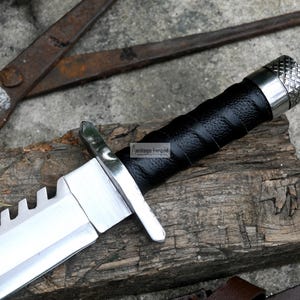 Pode incluir: Uma faca de ca&ccedil;a prateada e preta com uma l&acirc;mina serrilhada e um cabo de couro preto. A faca tem um pomo de metal com um design texturizado.