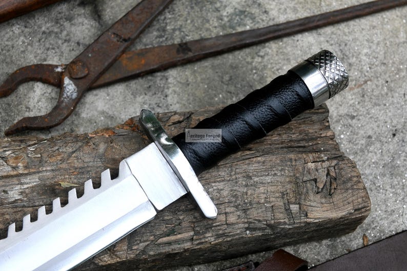 Pode incluir: Uma faca de ca&ccedil;a prateada e preta com uma l&acirc;mina serrilhada e um cabo de couro preto. A faca tem um pomo de metal com um design texturizado.