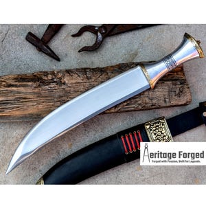 May include: A silver and gold curved dagger with a black leather sheath. The dagger has a decorative handle with gold accents and a sharp blade. The sheath has a gold accent and red stitching. The text "Heritage Forged" is visible on the sheath.  Forged with Passion, Built for Legends.
