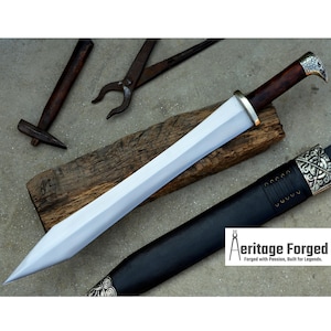 May include: A silver and brown sword with a leather scabbard. The sword has a sharp point and a double-edged blade. The scabbard has a decorative metal tip and a strap. The sword is resting on a wooden surface. The text "Heritage Forged" is visible in the lower right corner of the image.  Forged with Passion, Built for Legends.
