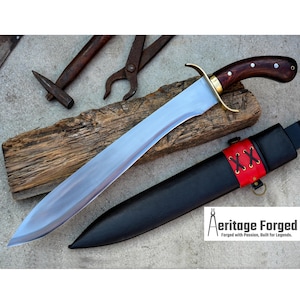May include: A long, silver-colored sword with a brown wooden handle and a black leather sheath. The sword is resting on a wooden surface with a hammer and pliers in the background. The text "Heritage Forged" is visible in the lower right corner of the image.