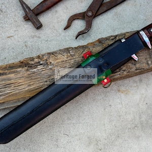 May include: A black leather sheath with red and green accents, holding a knife with a brown wooden handle and a silver blade. The sheath is resting on a piece of wood. The text "Heritage Forged" is visible on the sheath.