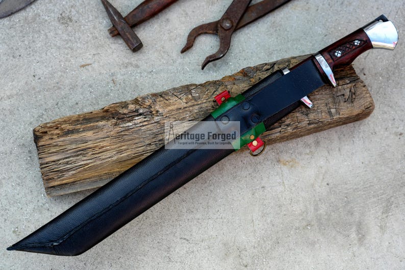 May include: A black leather sheath with red and green accents, holding a knife with a brown wooden handle and a silver blade. The sheath is resting on a piece of wood. The text "Heritage Forged" is visible on the sheath.