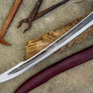 May include: A medieval-style sword with a silver blade and a decorative hilt, resting on a wooden plank. A burgundy leather sheath with green accents is visible. Also, a hammer and pliers are in the image.