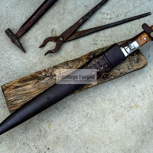 May include: A long, brown leather sheath with a wooden handle and a silver blade. The sheath is secured with a leather strap and has a metal buckle. The blade is sharp and pointed. The sheath is resting on a piece of wood. The text "Heritage Forged" is visible on the sheath.