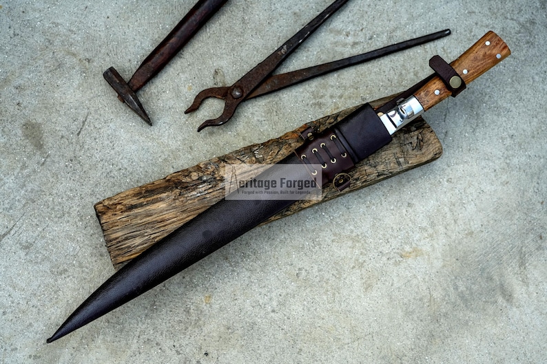 May include: A long, brown leather sheath with a wooden handle and a silver blade. The sheath is secured with a leather strap and has a metal buckle. The blade is sharp and pointed. The sheath is resting on a piece of wood. The text "Heritage Forged" is visible on the sheath.