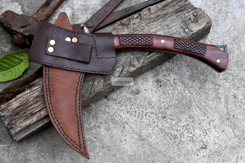 May include: A handcrafted hatchet with a dark wooden handle and a brown leather sheath. The hatchet head is made of metal and has a sharp edge. The handle features a textured grip for secure handling. The sheath is secured with brass rivets.