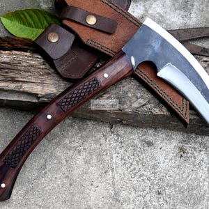 May include: A handcrafted axe with a curved blade and a dark wooden handle. The handle features a diamond pattern and is accented with small white rivets. The axe is accompanied by a brown leather sheath and rests on a weathered wooden surface.