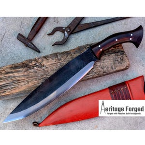 May include: A large, black, single-edged knife with a wooden handle. The knife is resting on a piece of wood and has a red leather sheath with brown straps. The text "Heritage Forged" is visible in the lower right corner of the image.  Forged with Passion, Built for Legends.