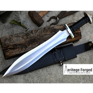Puede incluir: Una espada de acero pulido con empuñadura de cuero negro y funda a juego. La hoja tiene una forma curva distintiva. El texto "Aeritage Forged" es visible en la funda. La espada se exhibe sobre una tabla de madera.