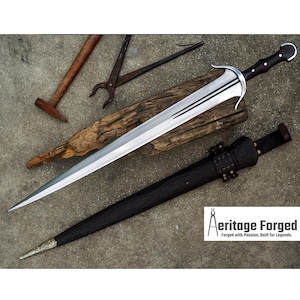 Könnte beinhalten: Ein silbernes und schwarzes Schwert mit gebogener Klinge, das auf einem Holzstück liegt. Eine schwarze Scheide und Werkzeuge sind ebenfalls vorhanden. Das Schwert hat einen schwarzen Griff und einen silbernen Parier. Der Text "Aeritage Forged" ist sichtbar.