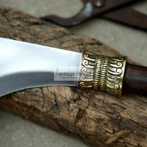 May include: A curved, silver blade with a wooden handle and a decorative brass pommel. The pommel has an intricate design of swirling lines and patterns.