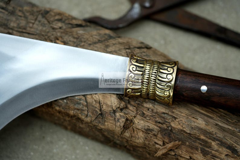 May include: A curved, silver blade with a wooden handle and a decorative brass pommel. The pommel has an intricate design of swirling lines and patterns.