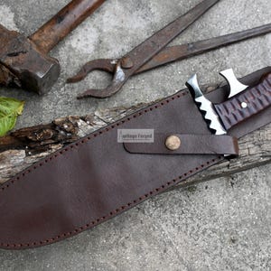 Puede incluir: Una funda de cuero marr&oacute;n con un borde cosido y un bot&oacute;n de metal. Un cuchillo con un mango de madera marr&oacute;n y una hoja plateada est&aacute; dentro de la funda. La funda est&aacute; sobre un trozo de madera.